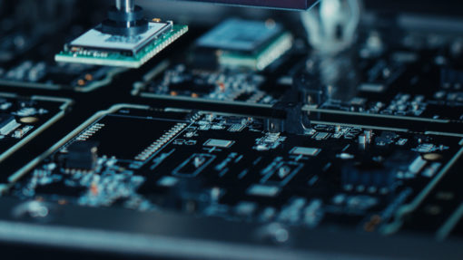 This is a detailed image of a computer motherboard, predominantly black in color. The focus is on the center where two green microchips are visible. To the right, there’s a silver robotic arm. Various other components like capacitors and transistors are scattered across the board. The background is blurred, hinting at more components and wires.