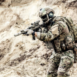 This is a photo-realistic image of a soldier in full gear, aiming a rifle in a desert-like environment. The soldier is equipped with a helmet with a visor and a camouflage uniform. The rifle is fitted with a scope and a suppressor. The background reveals a rocky terrain with loose soil and boulders. The image has an overall sepia tone, enhancing the desert ambiance.