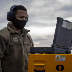 The image features a person wearing a gray hoodie and black headphones, standing next to a yellow and black equipment case. The equipment case is strikingly yellow with black stripes and bears a warning label. The backdrop is a cloudy sky, adding to the overall ambiance.