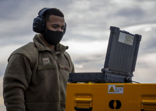 The image features a person wearing a gray hoodie and black headphones, standing next to a yellow and black equipment case. The equipment case is strikingly yellow with black stripes and bears a warning label. The backdrop is a cloudy sky, adding to the overall ambiance.