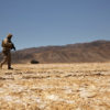 This is a photo-realistic image of a soldier walking alone in a barren desert. Dressed in a camouflage uniform and carrying a rifle, the soldier traverses the cracked soil of the desert. In the distance, mountains rise against a clear, blue sky. The image conveys a profound sense of solitude and desolation.