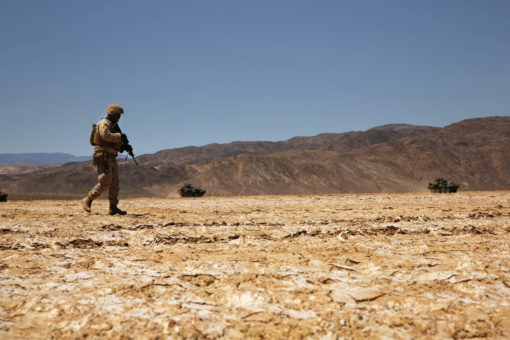 This is a photo-realistic image of a soldier walking alone in a barren desert. Dressed in a camouflage uniform and carrying a rifle, the soldier traverses the cracked soil of the desert. In the distance, mountains rise against a clear, blue sky. The image conveys a profound sense of solitude and desolation.