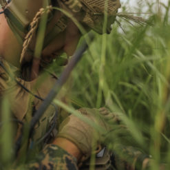 This photorealistic image depicts a soldier, clad in a camouflage uniform and helmet with a netted cover, lying in tall, green grass. The soldier is holding a rifle, ready for action. The image is cropped to show only the soldier’s upper body, adding to the tense mood of the scene. The setting appears to be a field or meadow, suggesting the soldier is on a mission or engaged in combat.