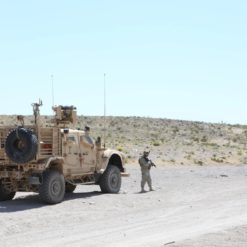 The image features a military vehicle in a desert setting. The vehicle, painted in a tan color to blend with the environment, is equipped with a large antenna, suggesting communication capabilities. It is parked on a dirt road, with a person standing nearby. The surrounding landscape is barren, dotted with rocks and sparse shrubs typical of a desert. Above, the sky is clear and blue, devoid of clouds.