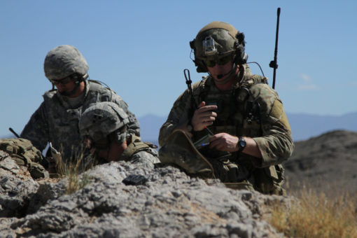 This photorealistic image portrays three soldiers in full gear stationed on a rocky hilltop. Dressed in camouflage clothing and helmets, they are prepared for action. The soldier on the right is holding a radio, while the one on the left wields a rifle. The backdrop is a clear blue sky and a rugged, mountainous landscape, adding to the intensity of the scene.