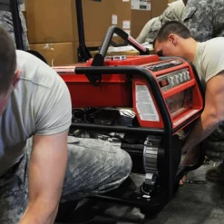 The image captures two unidentified individuals in U.S. military uniforms, diligently working on a red Honda EU3000iS generator. They are kneeling on the ground, engrossed in their task. The setting appears to be a warehouse or storage area, with boxes and other equipment visible in the background.