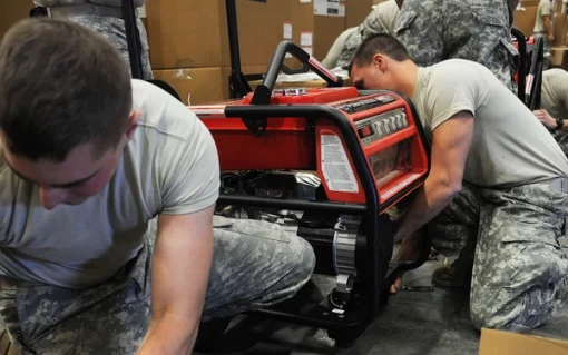 The image captures two unidentified individuals in U.S. military uniforms, diligently working on a red Honda EU3000iS generator. They are kneeling on the ground, engrossed in their task. The setting appears to be a warehouse or storage area, with boxes and other equipment visible in the background.