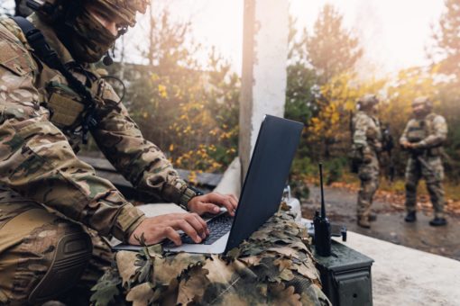 This image portrays a person in camouflage attire, sitting cross-legged on a rock and using a black, modern laptop in an outdoor setting. The individual is equipped with a backpack and has a rifle slung over their shoulder, suggesting preparedness for combat. In the background, trees with yellow leaves can be seen, along with another person in camouflage clothing standing at a distance. The scene appears to be set during daylight hours, highlighting the blend of military readiness and digital technology.