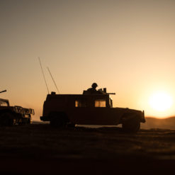 This image depicts two toy military vehicles, a jeep and a truck, both equipped with mounted guns, positioned on a sandy surface. The vehicles are silhouetted against a large, orange setting sun on the horizon. The sky transitions from orange to yellow, creating a warm, nostalgic atmosphere. The sand is light brown with slight ripples, adding to the realistic portrayal of a desert environment.