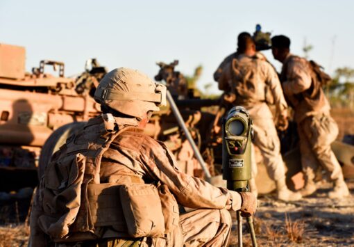 A U.S. soldier, donned in camouflaged combat gear and a sand-colored helmet, sits facing away from the viewer. He is closely monitoring a green, cylindrical-shaped optical device, possibly for surveillance or targeting, set on a tripod. To his right, the blurred forms of several other soldiers are seen engaged in activities beside a desert-tan armored vehicle with complex equipment on top. The background reveals an arid landscape bathed in the soft light of either dawn or dusk. The atmosphere suggests a moment of intense focus during a military operation.