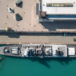 An aerial view of a large naval vessel moored alongside a rectangular pier. The ship, painted in grey, displays a combination of technical equipment on its deck, including radar systems, weapon mounts, and helicopter landing pads. Adjacent to the ship, the pier features scattered individuals, possibly naval personnel, walking or standing, and various structures and storage items. Notably, a long, cylindrical object, perhaps a protective covering or equipment, lies on one side of the pier. The water surrounding the vessel is a vibrant turquoise, and a small tugboat with trailing ropes is situated near the ship's bow, possibly assisting with mooring or refueling operations.