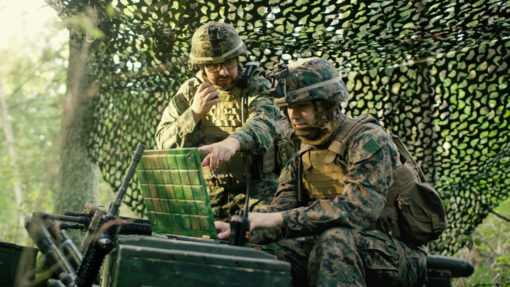 Two soldiers in full combat gear, nestled under a camouflage net in a dense forest. The soldier on the left speaks into a radio while the one on the right points at a green, grid-like digital device. In the foreground, a mounted assault rifle rests against an ammunition box. Sunlight filters through the net and surrounding trees, casting dappled light on their focused expressions. Their surroundings indicate a covert or strategic operation in a natural environment.