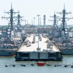 This image shows two large U.S. navy ships docked at a concrete pier. The ships are grey in color and feature multiple levels and antennas. A red buoy is floating in the light blue-green water in front of the pier. In the background, a city skyline under a blue sky can be seen.