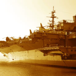 This is a sepia-toned image of a large U.S. aircraft carrier sailing on the ocean. The carrier has multiple levels, including a flight deck where several aircraft are parked. The control tower and other structures are visible on the carrier. The horizon can be seen in the background, indicating that the image was taken from the side of the carrier.