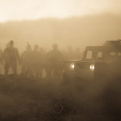 A sepia-toned image captures a group of soldiers and a military vehicle advancing in a foggy or smoky environment. The soldiers, walking in a line, follow the vehicle which is equipped with a mounted gun and has its headlights on. The figures are blurry, adding to the mysterious atmosphere. The hazy backdrop reveals the faint outlines of trees and hills.