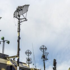 The image provides a low-angle view of a military vehicle equipped with an array of antennas and satellite dishes, mounted on poles and pointing in various directions. The vehicle, painted in a green camouflage pattern, stands out against the backdrop of a blue sky with scattered clouds. The antennas and dishes, rendered in black and gray, have a metallic appearance, reflecting their robust construction. This setup suggests a sophisticated communication system, vital for coordinating military operations.