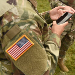 The image depicts a U.S. soldier in a camouflage uniform, holding a black mobile phone with both hands. The soldier’s uniform displays an American flag patch on the left arm, signifying their service to the country. The setting is a grassy outdoor area, and another individual in a military uniform can be seen in the background, suggesting a field operation or training exercise.