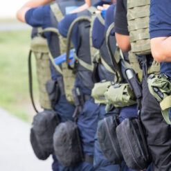 The image showcases a group of individuals dressed in navy blue uniforms, standing in a line and facing the same direction, suggesting a formation or parade. They are equipped with green and black backpacks that feature multiple pockets and straps, indicating readiness for a mission or travel. The setting is an outdoor grassy area with a sidewalk visible in the background. One individual is notably carrying a gun in a holster on their hip, further emphasizing the military context. The image is cropped to show only the torsos and legs of the personnel.