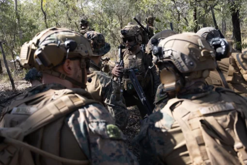 A group of U.S. military soldiers in full gear are conducting an operation in a wooded area. They are standing in a circle, facing each other, suggesting a strategic discussion. Each soldier is equipped with a helmet that has night vision goggles attached, indicating readiness for nocturnal activities. Their camouflage uniforms blend with the surrounding trees and shrubs, providing them with a tactical advantage. They are also carrying rifles, prepared for potential combat situations. One soldier’s face is intentionally blurred out, respecting privacy and confidentiality norms.”