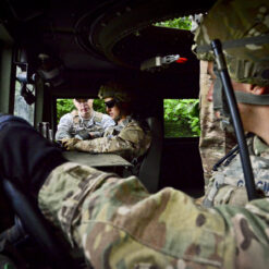 This image provides a first-person perspective from the driver’s seat of a U.S. military vehicle stationed in a wooded area. The driver, dressed in a camouflage uniform and a helmet equipped with a microphone, has both hands on the steering wheel, ready for action. The passenger seat and back seat are occupied by personnel whose faces are respectfully blurred for privacy. An open window on the passenger side lets in the ambient sounds of the forest. Adding a touch of patriotism, a red and white flag hangs from the rearview mirror, symbolizing the spirit of the mission.