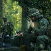 This image depicts a U.S. military operation in a forest setting. The main focus is a soldier, fully dressed in camouflage gear, sitting at a desk equipped with a laptop and a radio. The soldier, wearing a helmet with an attached headset, is actively communicating via the radio. The desk, covered with camouflage netting, holds a large black case, suggesting important equipment or documents. In the background, two additional soldiers in full camouflage gear can be seen amidst the trees and foliage, further emphasizing the covert nature of the operation. Privacy is maintained with blurred faces.