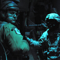 Pilot in green flight suit and helmet illuminated by dim blue light inside an aircraft, facing two camouflaged soldiers. The pilot's patch, and the detailed gear of the soldiers are subtly visible in the low-light environment.