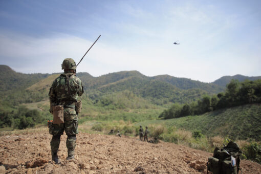 Soldier in camouflage uniform stands on a rugged terrain, holding a long radio antenna, looking out towards rolling hills. In the distant sky, a helicopter is visible. Further down the terrain, a group of soldiers gather, and a packed military backpack sits in the foreground