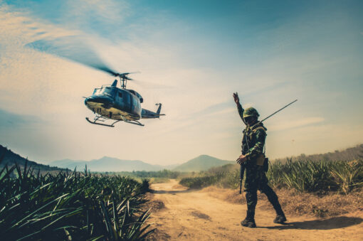 "Soldier in full gear signals with raised hand to a hovering helicopter against a clear sky. Below, a rugged path cuts through tall grasses, leading to distant mountains."