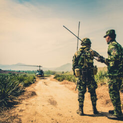 Two soldiers, dressed in camouflage uniforms and helmets, stand on a dusty path surrounded by tall, wild grass. The soldier on the left holds a long radio antenna, indicative of a communication role, while the one on the right seems to be giving instructions. Further down the path, a helicopter with its blades still, waits on a makeshift landing zone. The backdrop reveals rolling hills under a vast sky, suggesting a remote or rural location. The overall scene captures a moment of coordination and readiness during a field operation.
