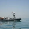 A sleek, gray military patrol boat equipped with advanced navigation and communication systems floats on calm, clear waters. The boat, characterized by its inflatable side borders, has an American flag flying on its aft end. Mounted on the boat is specialized equipment, including a small orange lifeboat. In the distance, a faint coastline can be seen against a clear blue sky.