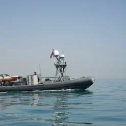 A sleek, gray military patrol boat equipped with advanced navigation and communication systems floats on calm, clear waters. The boat, characterized by its inflatable side borders, has an American flag flying on its aft end. Mounted on the boat is specialized equipment, including a small orange lifeboat. In the distance, a faint coastline can be seen against a clear blue sky.