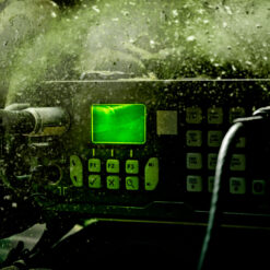 A close-up view of a rugged military communication console, illuminated predominantly by a green hue. The console has a bright, rectangular green screen displaying wave patterns. Below the screen, there are tactile buttons labeled with various functions such as "F1", "F2", "F3", and symbols like "X" and "Q". The surrounding area is dusty and weathered, suggesting wear from use in rough conditions. In the foreground, parts of two wrapped cables can be seen, one notably thicker than the other. The overall impression is of a durable device designed for crucial communications in challenging environments.