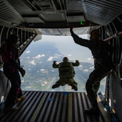 Inside the spacious cargo bay of a military aircraft, three individuals prepare for a skydive. The scene is backlit, emphasizing their silhouettes. On the left, a jumper in a black and pink jumpsuit stands tensely by the edge. In the center, another jumper, dressed in green military gear, is captured mid-jump as he leaps outward, arms and legs spread. To the right, another individual in military attire grips an overhead handle, watching the jumper. Below, a vast landscape of green fields, roads, and scattered clouds stretches out, emphasizing the great altitude.