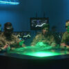 "Four U.S. military personnel in a high-tech command center, wearing green-glowing augmented reality goggles, gather around a table emitting a holographic display. In the dim, blue-lit room, data-filled monitors and a tall server rack are visible in the background."